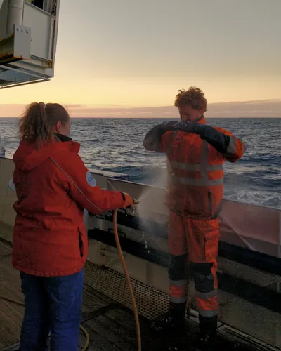 A woman directs a jet of water from a hose at a man on a ship's deck.