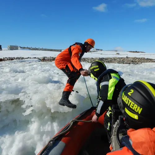 A man in a bright orange immersion suit climbs off an ice ledge into a small boat.