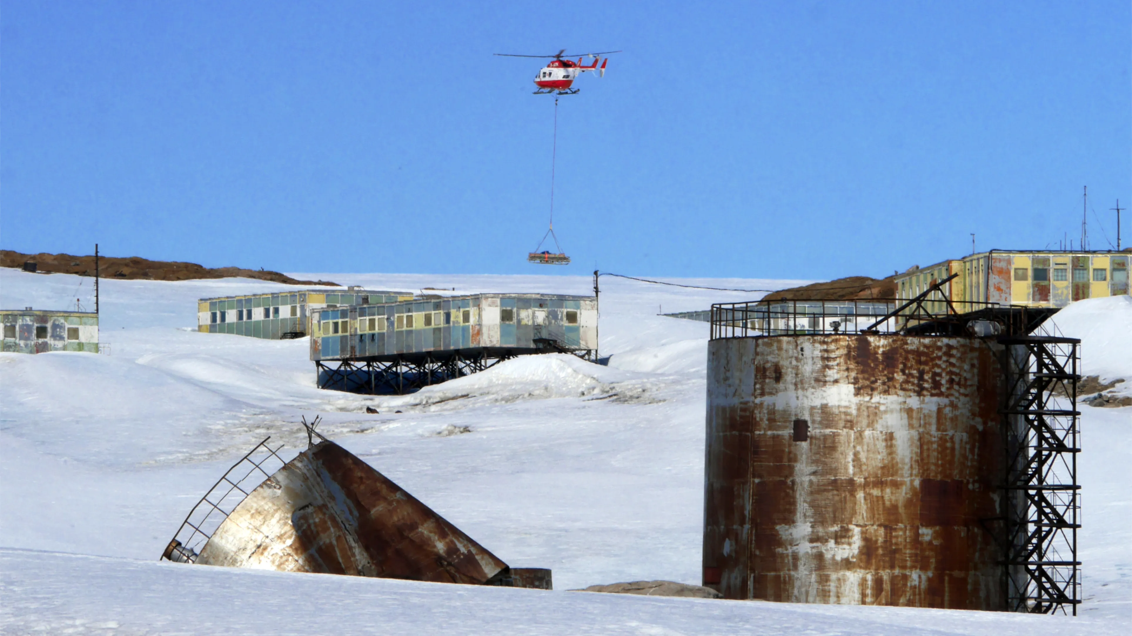 A red helicopter flies over a collection of old buildings on stilts in the snow with faded peeling paint and two metal silos in the foreground, one of which is crushed and broken.