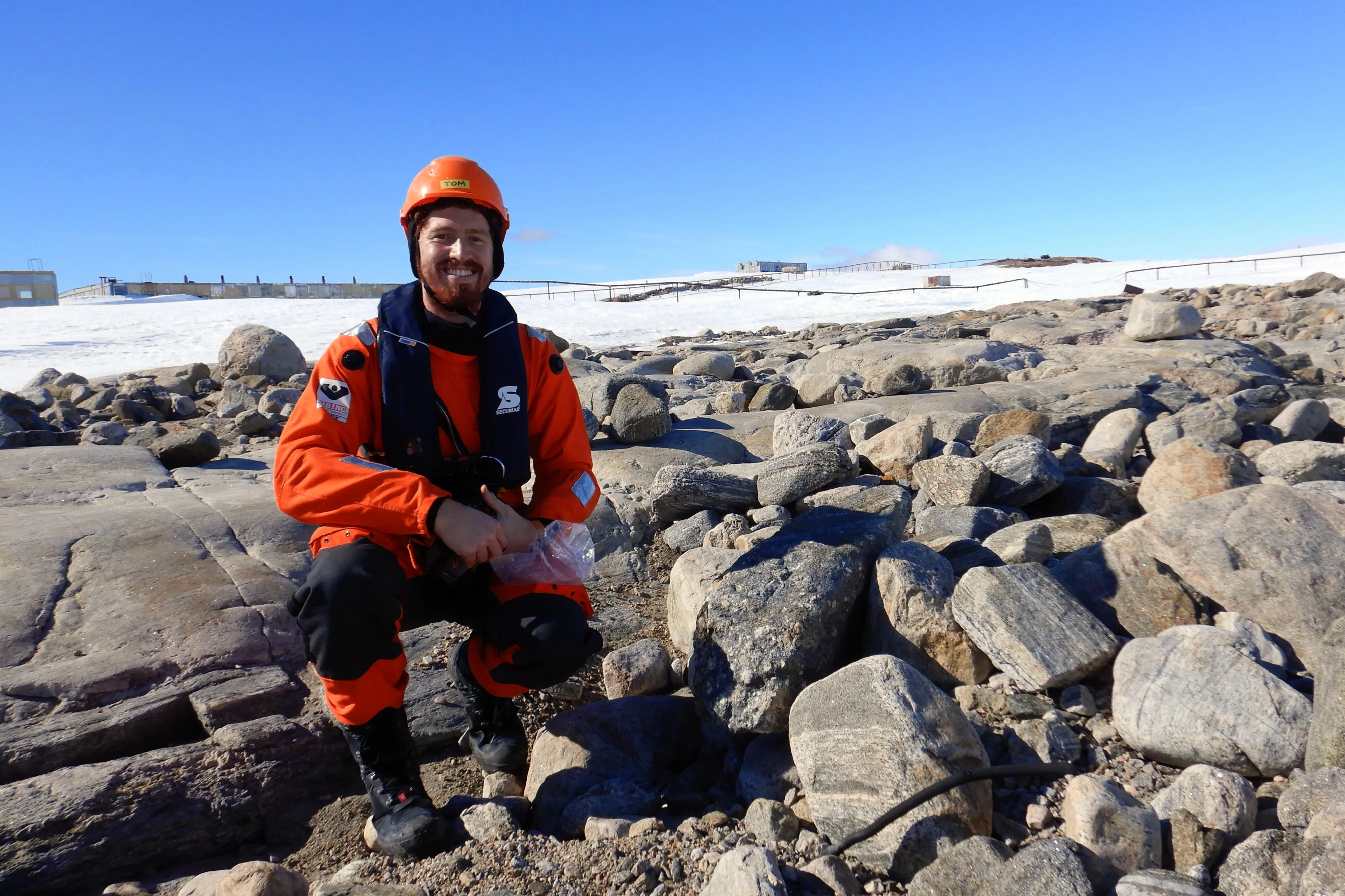 A man in a bright orange immersion suit squatting in a field of football-sized grey rocks.