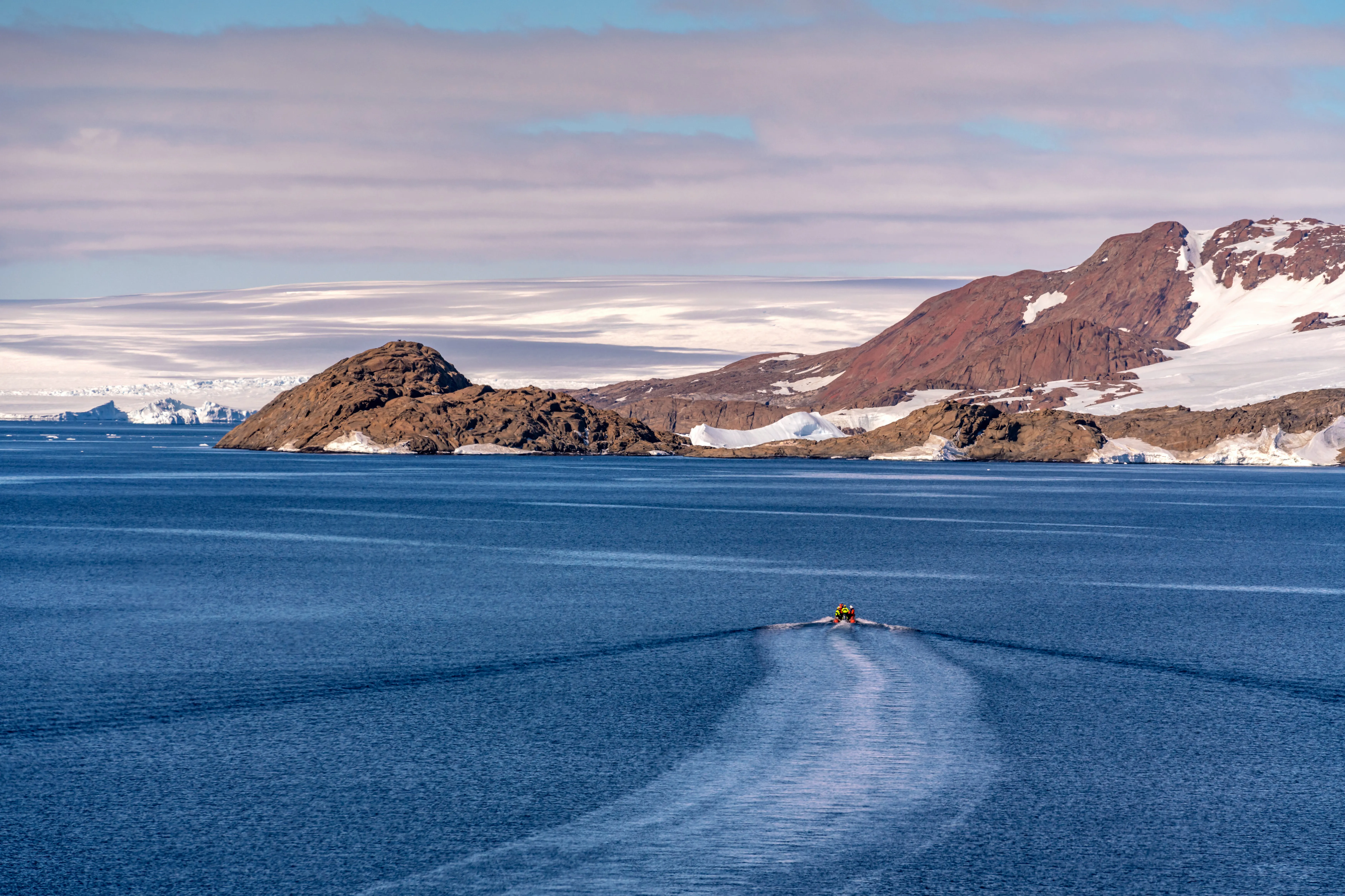 A small orange boat speeds off towards brownish-red snowy mountains which come right down to the sea.