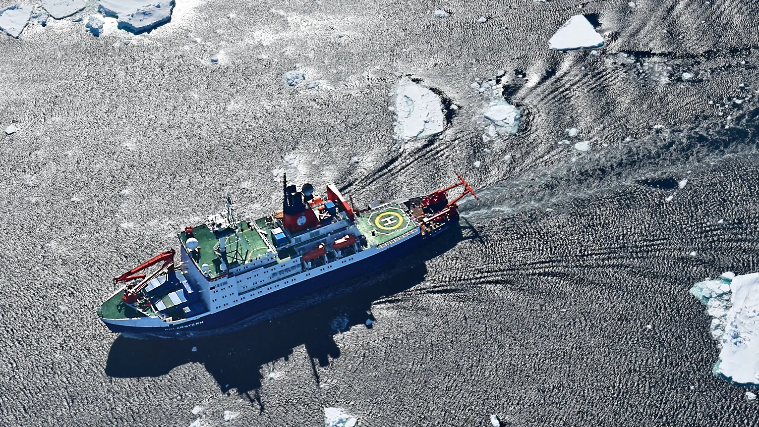 A blue and white ship seen from the air, steaming through a sea with scattered ice floes.