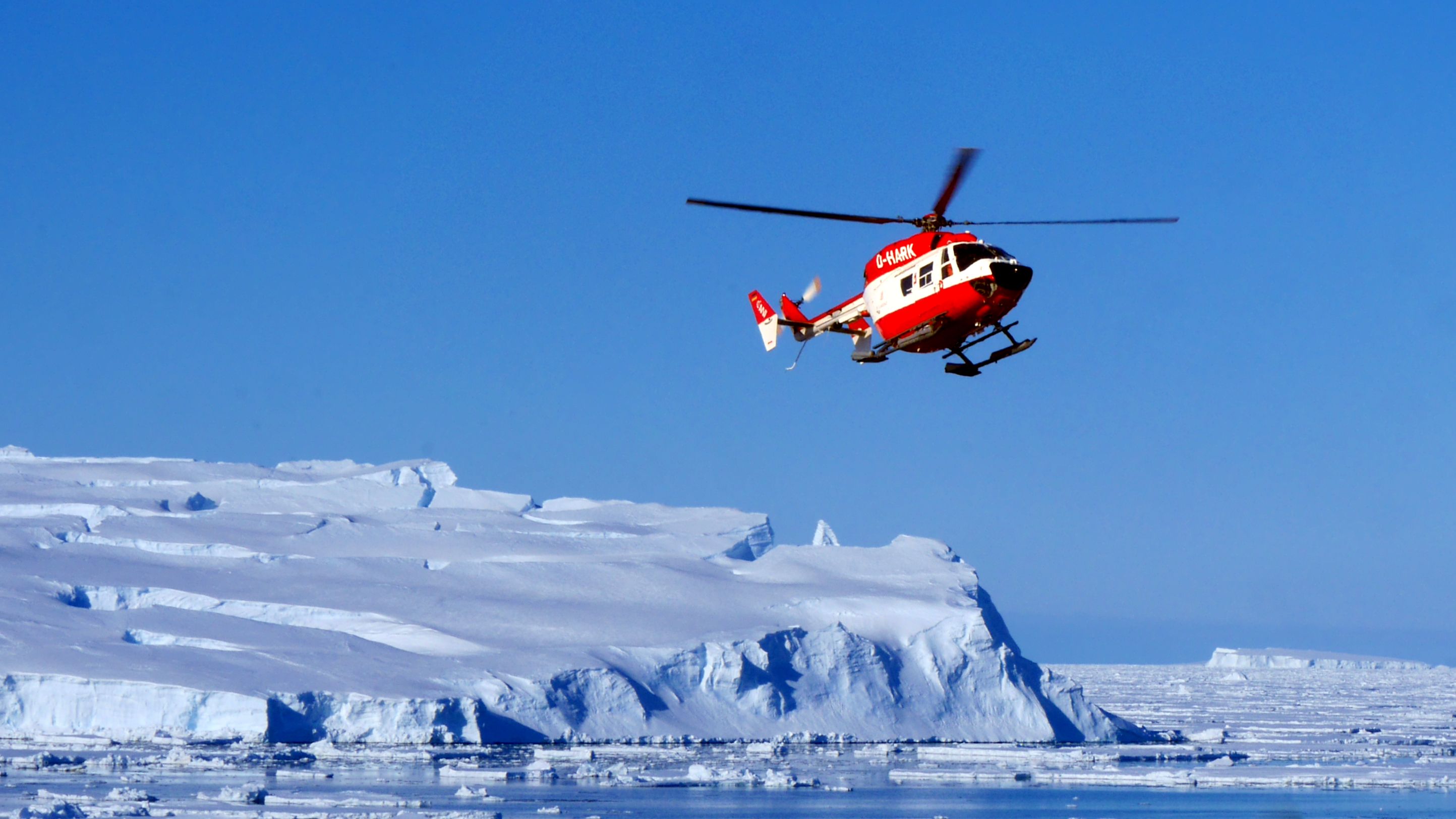 A red and white helicopter in clear blue skies in front of a large iceberg