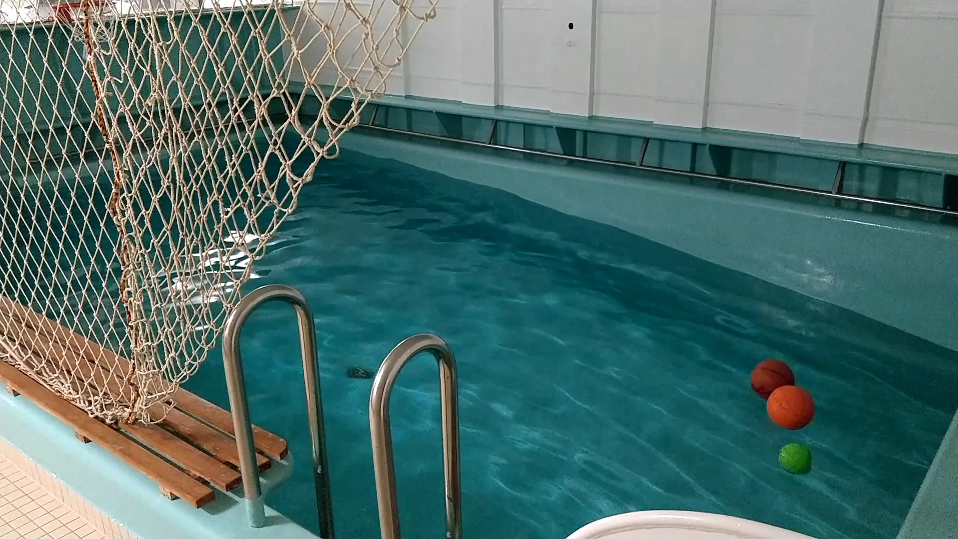 A light blue swimming pool with a basketball floating on the surface of the water, which is at a surprising angle.