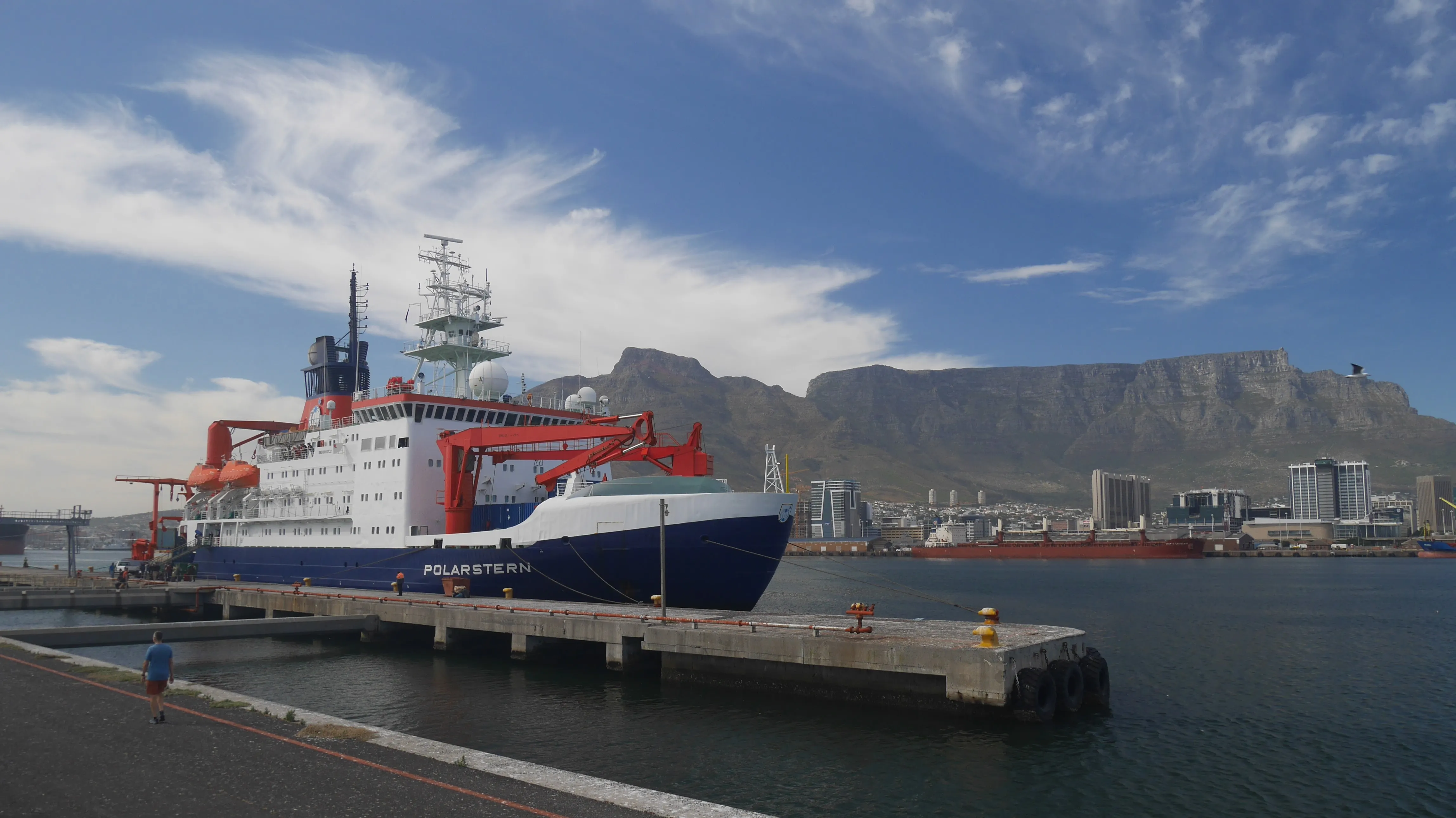 A blue and white ship in a harbour with a city in the middle ground and a mountain in the background