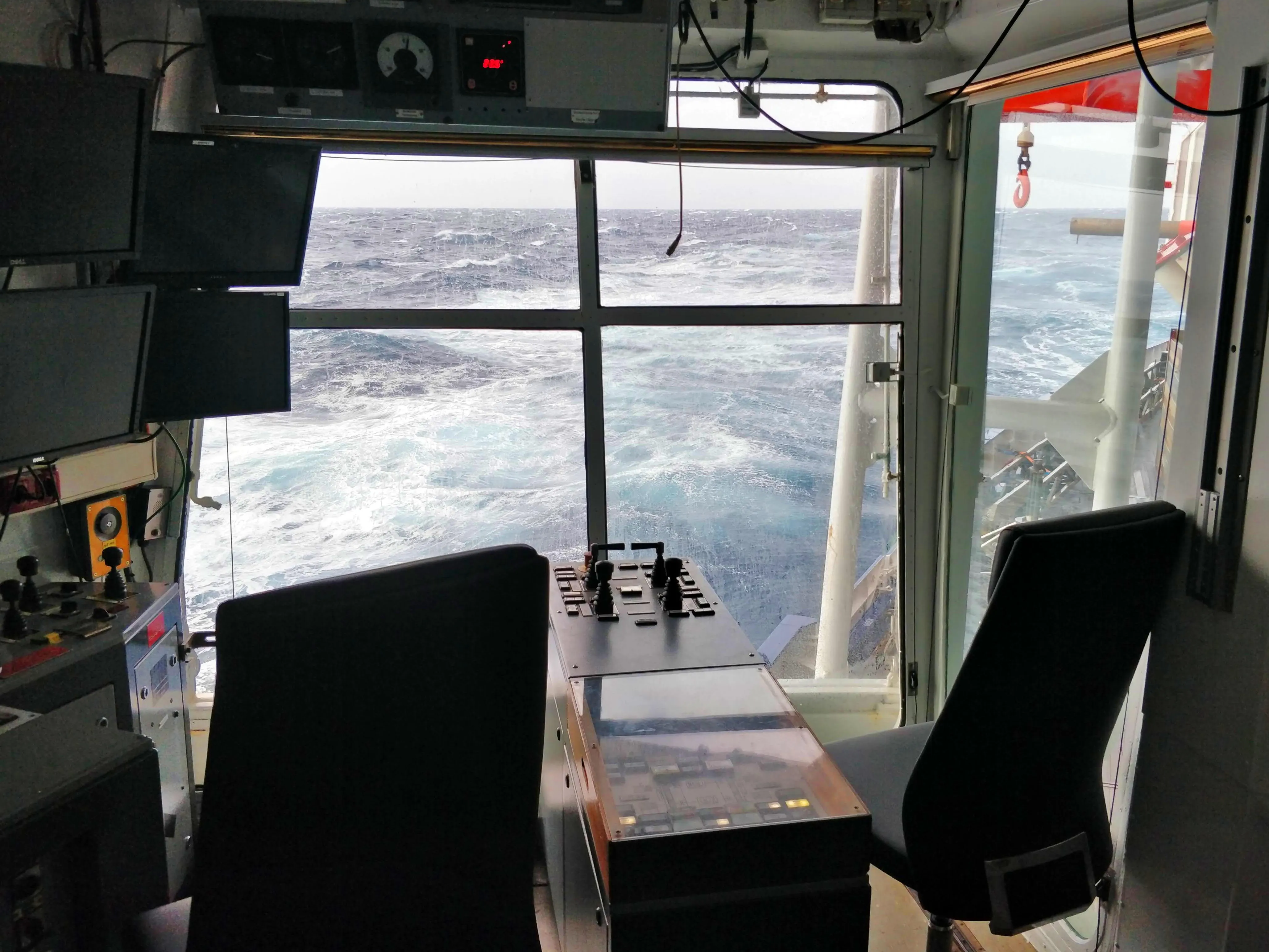 View of the choppy sea through the windows of a cabin with two seats and lots of screens and dials.