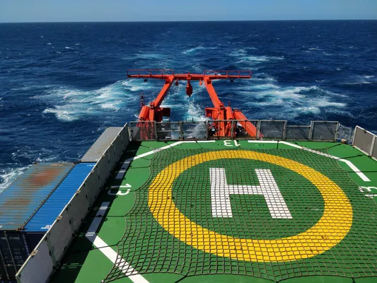 The open deck of a ship, painted green with a yellow circle and a white letter H. The wake of the ship extends away to the horizon in a deep blue sea.