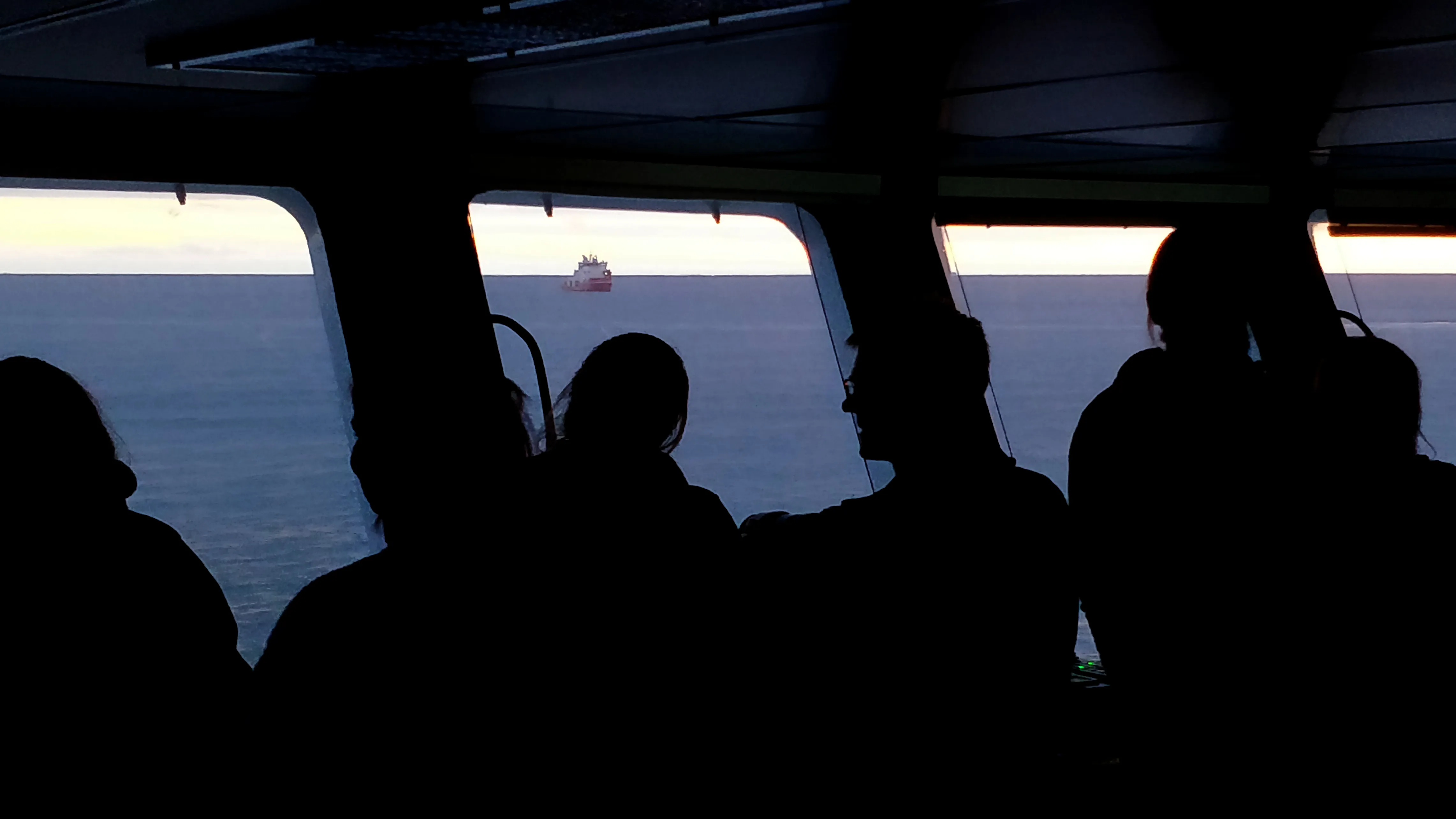 View of a red ship through the windows of a ships bridge. Many people are watching, all in silhouette against the windows.