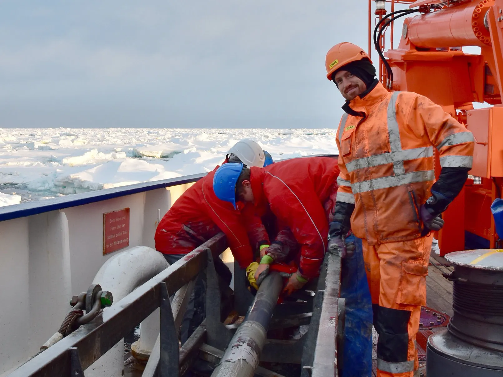 Two people are hard at work on a ship's working deck, while a man in muddy orange hi-vis clothing stands back and smiles at the camera.