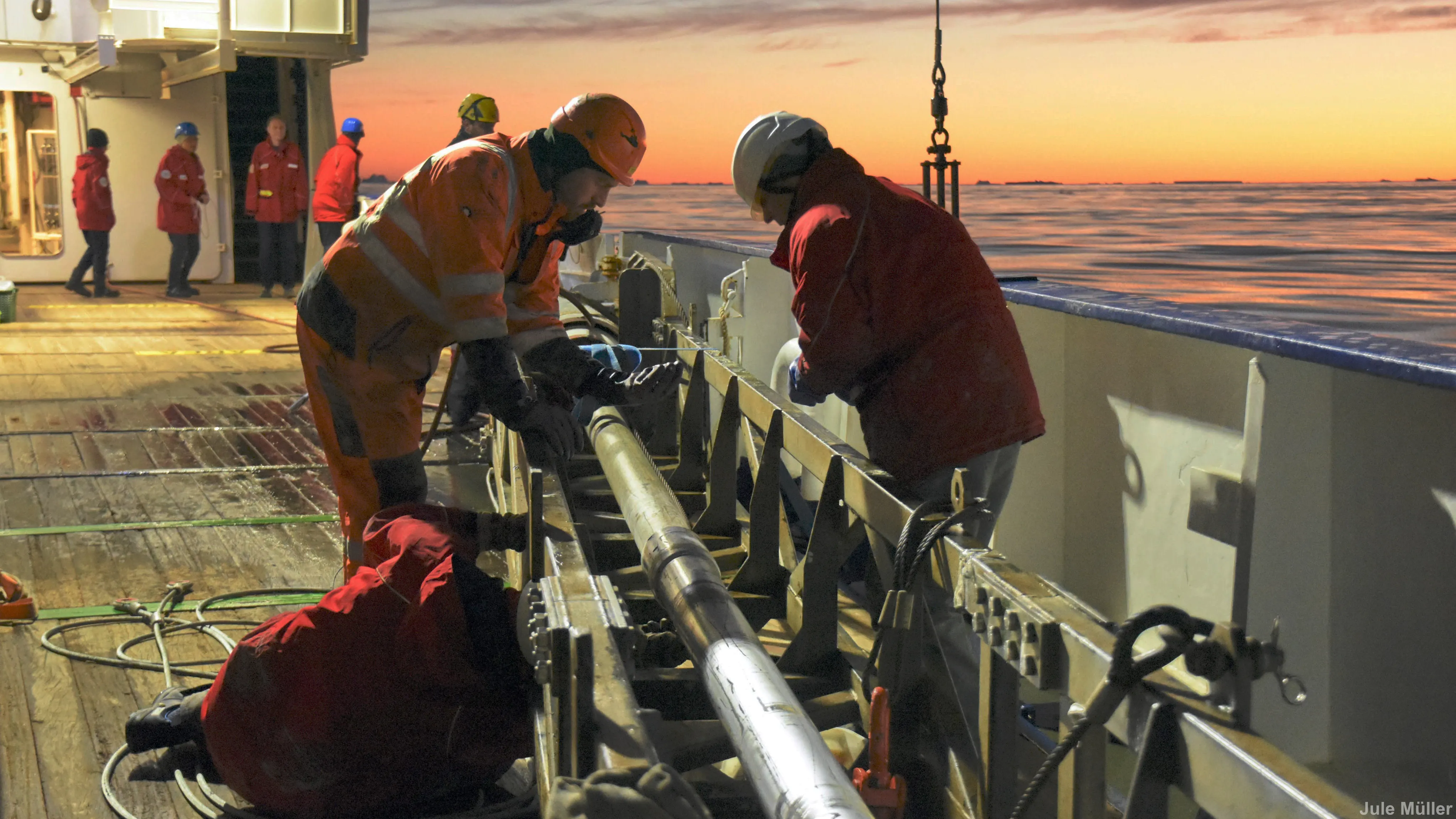 Three people working with what looks like a steel pipe in a metal support, on the deck of a ship at sunset.