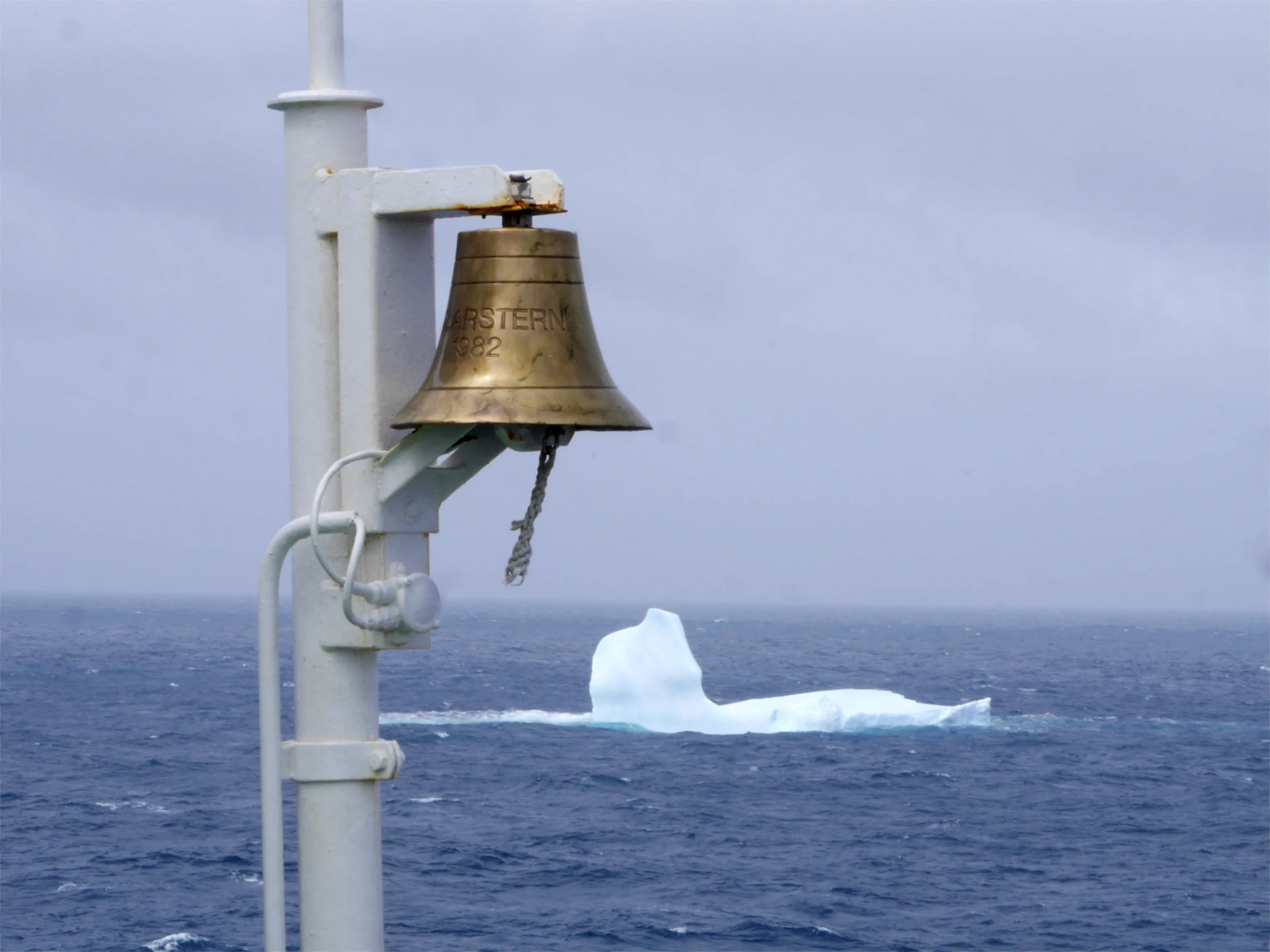Photo of an iceberg in a grey ocean with a bell in the foreground. The iceberg has the rough outline of a bird lying on its back, and the bell has 'Polarstern 1982' written on it.
