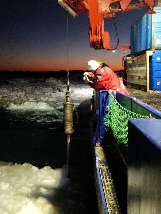 A steel pipe with a wider cylinder at the top hanging off a wire from a winch on a ship at sunset.