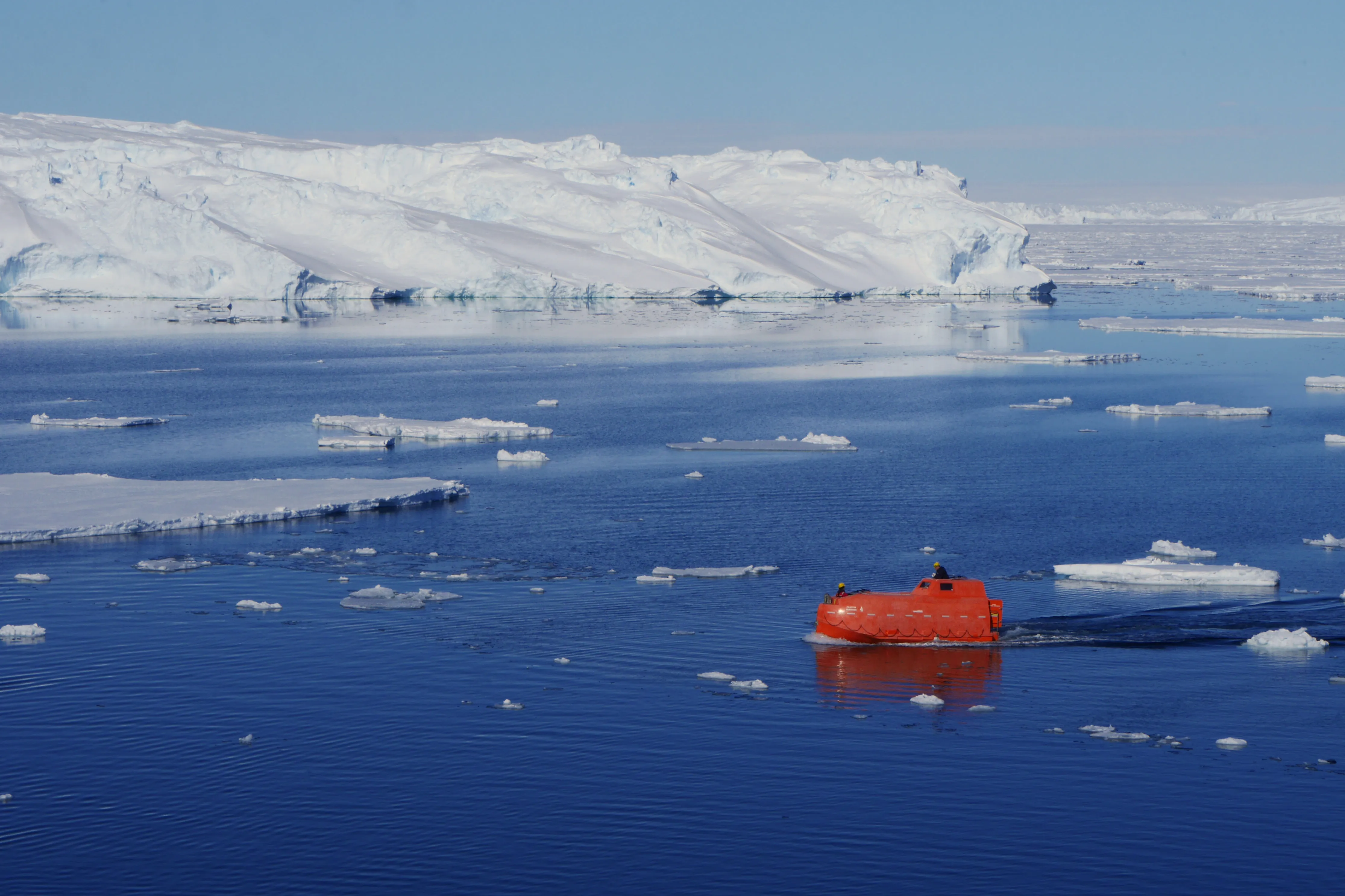 An orange ship's lifeboat motors across a still blue bay against a backdrop of bright white ice and snow.