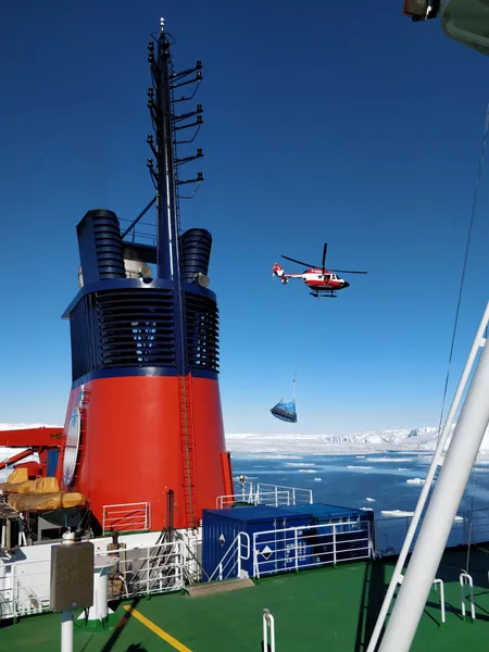 A helicopter carrying a load slung under it in a net on a cable passes the ship's funnel, headed towards the ice in the background.