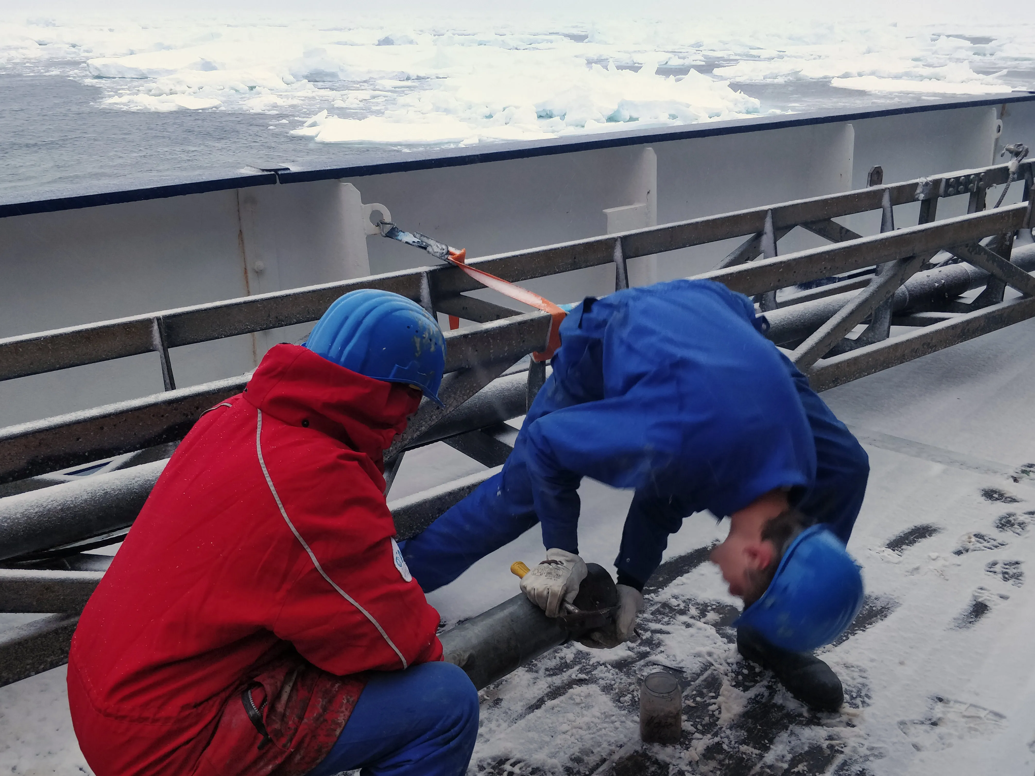 Two men working with what looks like a steel pipe on a snowy ship's deck, with sea ice in the background.