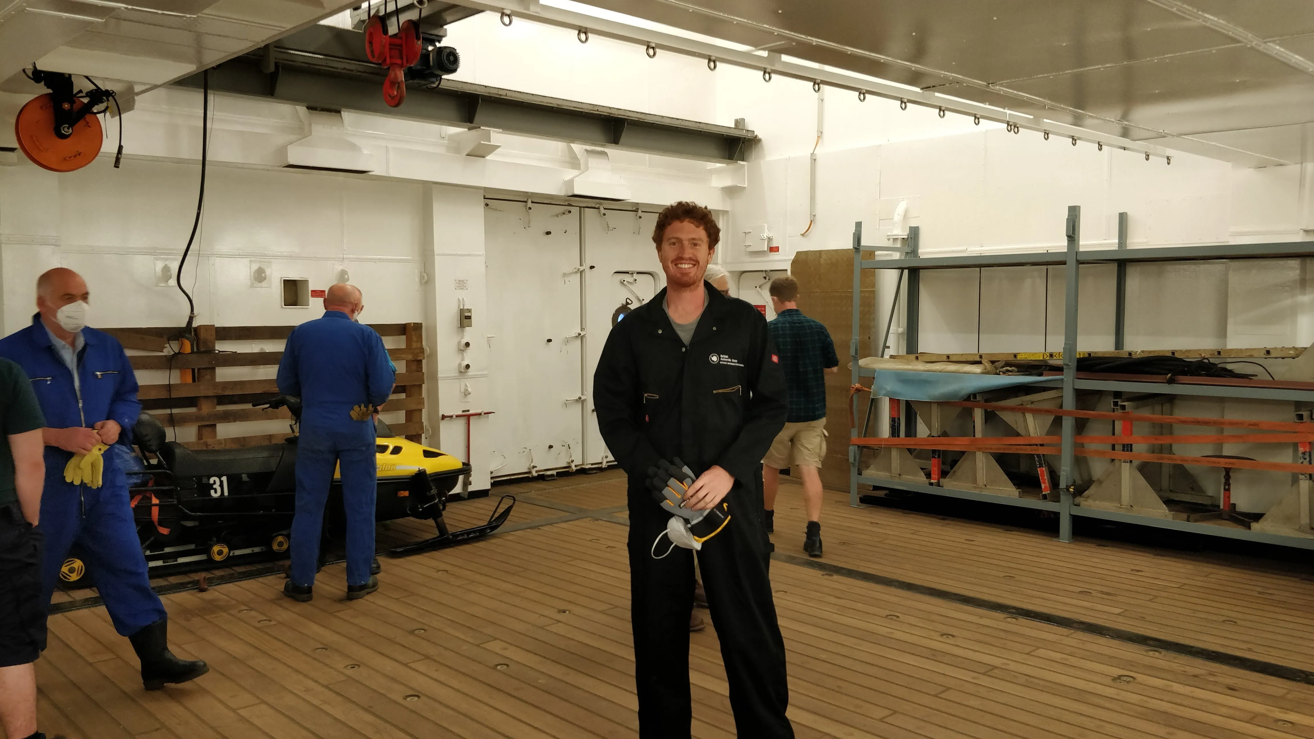 Photo of a man in a black boiler suit in a large space with wooden floor and white walls. Other people and some equipment are also in the room.